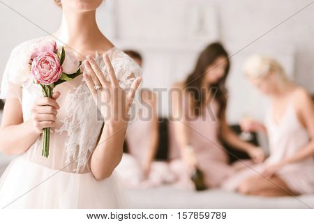 Engagement ring. Pleasant shy young bride standing in the white colored bedroom while showing her engagement ring and her bridesmaids having fun in the background