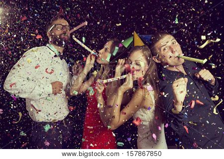 Two beautiful young couples having fun at New Year's party wearing party hats dancing and blowing party whistles. Focus on the couple on the right