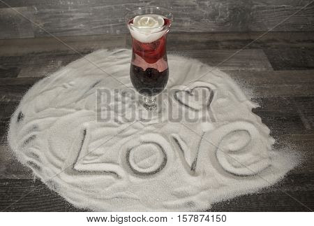 A cocktail glass with a floating rose blossom and a white candle in the sand. In the sand stands the word Love and a heart