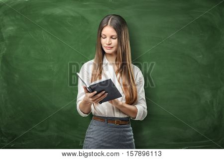 A young businesswoman holding her open day planner on the green chalkboard background. Daily planning. Time-management. Secretary work.