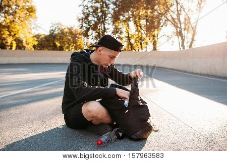 Young man looking for something in his sport bag. Cute guy sitting on stadium and rummaging in his backpack.