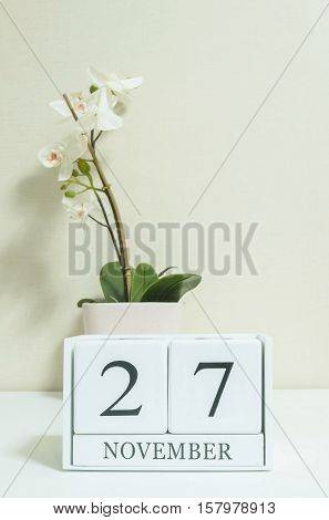 Closeup white wooden calendar with black 27 november word with white orchid flower on white wood desk and cream color wallpaper in room textured background selective focus at the calendar