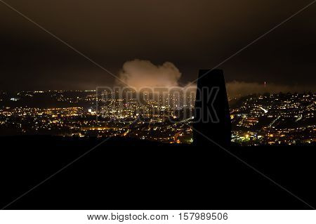 Silhouetted trig point on Little Solsbury Hill overlooking Bath. Night view of the UNESCO World Heritage City of Bath with stone used for cartographic triangulation in foreground