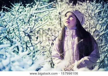 pretty preteen girl in knitted white hat and jacket on the snow bushes background under street light night scean
