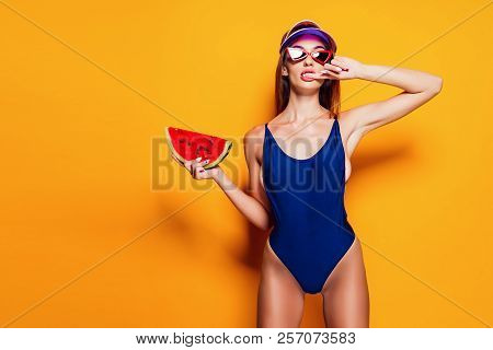 Beautiful Young Lady In Swimwear Biting Finger And Holding Piece Of Fresh Watermelon While Standing 