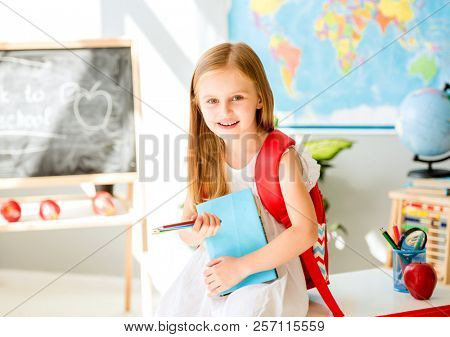 Little smiling blond girl standing near blackbord with red backpack and holding a notebook with pencils in the sunshine school classroom