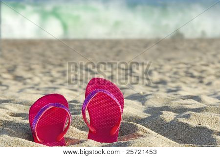 Flipflops สีชมพูในทรายกับคลื่นในระยะ