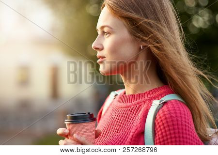 Large Portrait Of A Girl In A Sweater, With Coffee And A Backpack. The Woman Looks Into The Distance