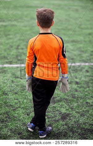 Boy goalkeeper stands in goal box on sports playground, rear view.