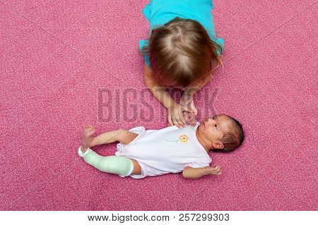 Bird Eye View Of An Older Sister Laying On Her Stomach, Holding The Hand Of Her Baby Sister To Comfo
