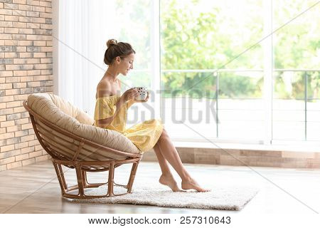 Young Woman With Cup Of Aromatic Coffee Sitting In Papasan Chair Near Window At Home