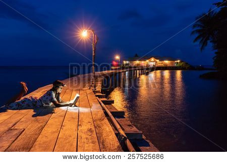Woman reading book at dusk, using forehead light, on a wooden bridge, Hatta island, Maluku, Indonesia
