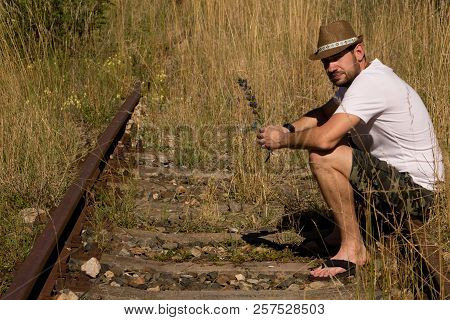 A Lonely Man In A Strawy Hat Sitting On A Rails And Holds A Flower In Hand.man Sitting On A Rails Ne