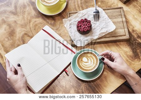 Cup Of Coffee Latte And Sweet Pastry On Wooden Table In Woman Hands. Hands Hold Opene Nove Book, Spr
