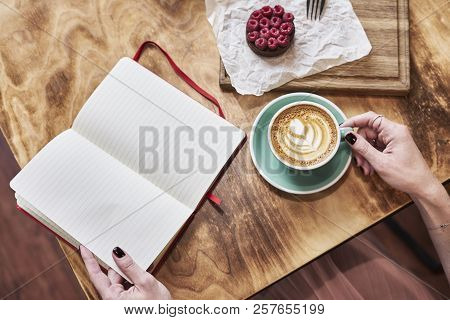 Cup Of Coffee Latte And Sweet Pastry On Wooden Table In Woman Hands. Hands Hold Opene Nove Book, Spr