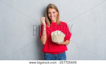 Beautiful young woman over grunge grey wall eating pop corn with a happy face standing and smiling with a confident smile showing teeth