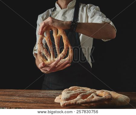 On a dark background and on a background of a wooden brown table, men's hands hold freshly baked bread fougas