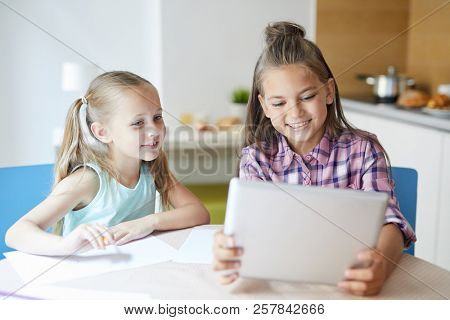 Cheerful cute little sisters watching curious vids or movie in tablet while spending weekend at home