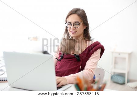 Young woman with baby in ergo carrier using laptop indoors