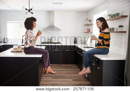 Two Female Friends Meeting For Coffee At Home Sitting On Kitchen Island
