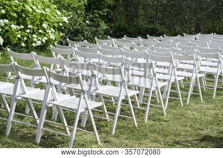 Empty Wooden Chairs In White, Arranged In Rows On The Lawn, The Background Of Hedges