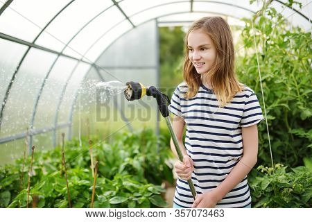 Adorable Young Girl Watering Variuos Vegetables In A Greenhouse On Sunny Summer Day. Gardening Activ