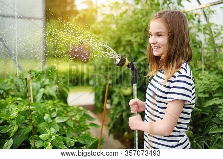 Adorable Young Girl Watering Variuos Vegetables In A Greenhouse On Sunny Summer Day. Gardening Activ