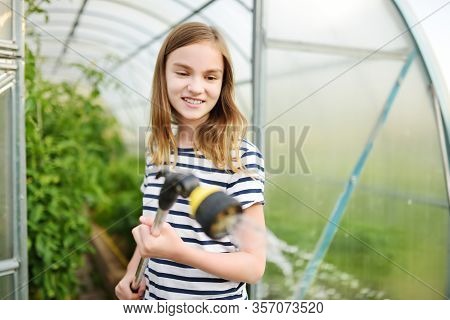 Adorable Young Girl Watering Variuos Vegetables In A Greenhouse On Sunny Summer Day. Gardening Activ