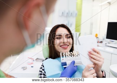 Patient In A Dental Office Holds A Palette With Shades Of Teeth And Looks In The Mirror