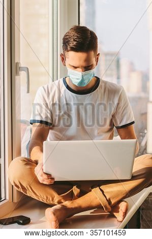 Young Man In Medical Protective Mask On Face Sitting On Windowsill With Laptop On Laps. Student Free