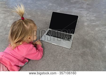 Small Cute Blond Caucasian Child Laying On Floor On Grey Rug And Watching Cartoons On Laptop. Black 