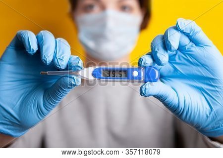 Woman In A Medical Mask With A High Fever. Holding A Thermometer In His Hands With A Reading Of 39.5