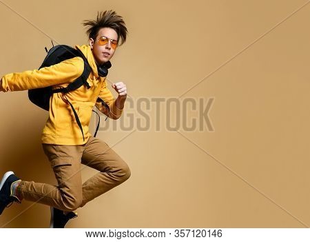 Young Teen Boy In Comfortable Clothing, Sneakers, Sunglasses And Backpack Jumping Over Yellow Wall B