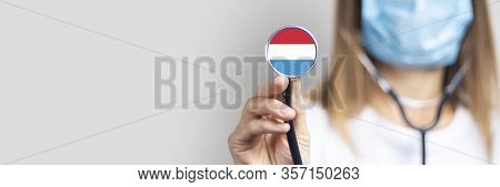 Female Doctor In A Medical Mask Holds A Stethoscope On A Light Background. Added Flag Of Luxembourg.