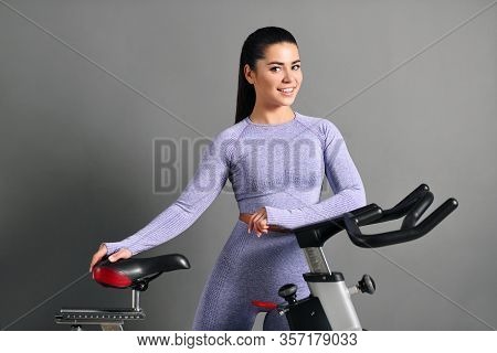 A Beautiful Athletic Young Brunette Woman In Sportswear Trains On A Sycle In The Gym Against The Gra