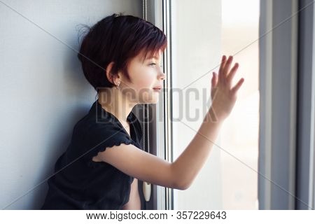 Sad Little Girl Looking Out Window Touching Glass By Hand. Child During Quarantine Because Of Pandem