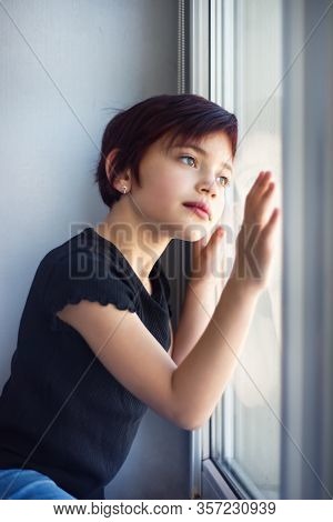 Vertical Picture Of Sad Little Girl Looking Out Window Touching Glass By Hand. Child During Quaranti