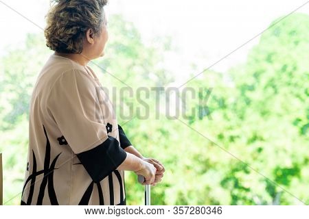 Elderly senior female using walking stick or walking cane staff standing beside window in domestic living room - recovery and rehabilitation concept