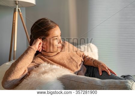 Staying home Asian woman relaxing pensive looking out the window sitting in living room sofa enjoying free time indoors during coronavirus outbreak.