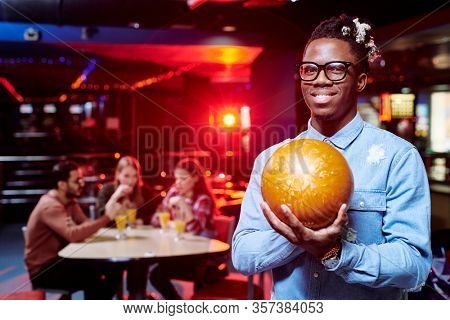 Happy young African man with bowling ball standing in front of camera during play on background of his friends having drinks