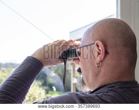 Elderly Man Sittiing, Looking Through A Glass Door With Binoculars As He Watches Something In The Di