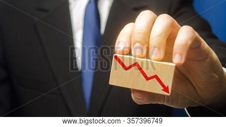 Businessman Holds Wooden Block With Red Down Arrow. The Concept Of Reducing Profits And Income. Rece
