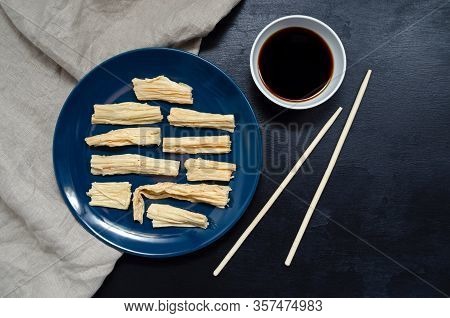 Yuba Tofu Skin On A Blue Plate On A Towel With Bamboo Sticks And Soy Sauce On A Black Slate Backgrou