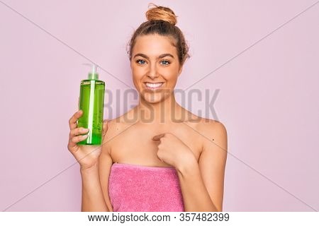 Beautiful blonde woman with blue eyes wearing towel shower after bath holding aloe vera gel with surprise face pointing finger to himself