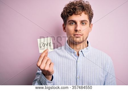 Young blond romantic man with curly hair holdiing paper with love message for valentines day with a confident expression on smart face thinking serious