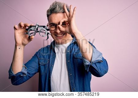 Young modern optical optomestrist man holding optometrical glasses over pink background with happy face smiling doing ok sign with hand on eye looking through fingers