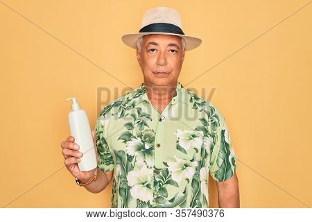 Middle age senior grey-haired man wearing summer hat holding sun protection cream with a confident expression on smart face thinking serious