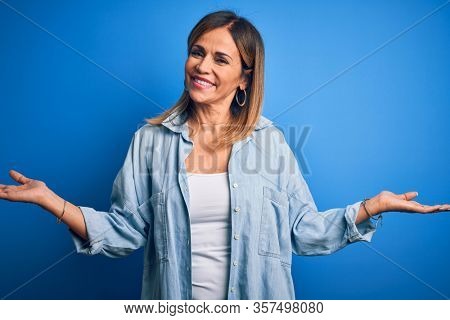 Middle age beautiful woman wearing casual shirt standing over isolated blue background smiling showing both hands open palms, presenting and advertising comparison and balance