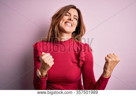 Young beautiful brunette woman wearing casual sweater stnding over pink background very happy and excited doing winner gesture with arms raised, smiling and screaming for success. Celebration concept.