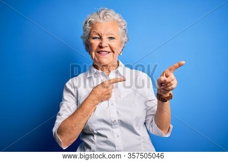Senior beautiful woman wearing elegant shirt standing over isolated blue background smiling and looking at the camera pointing with two hands and fingers to the side.